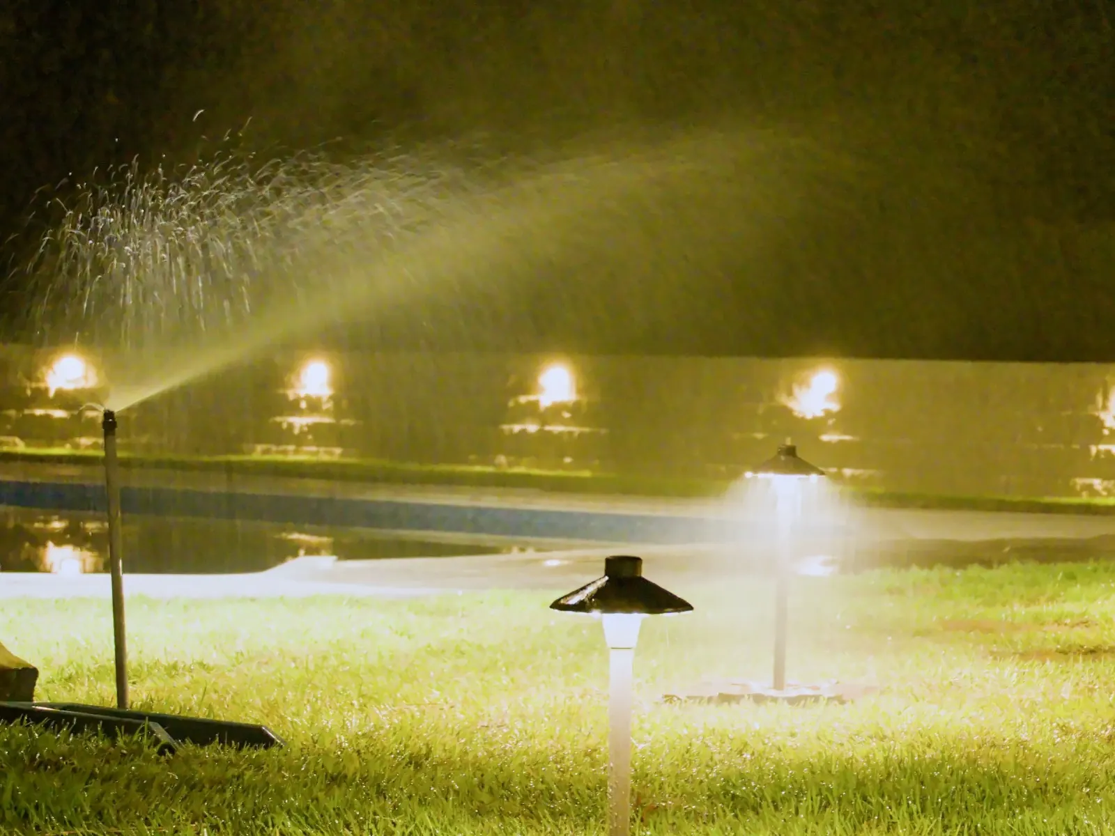 Irrigation head spraying at night across a lawn in Baldwin County, Alabama, with landscape lighting in the background