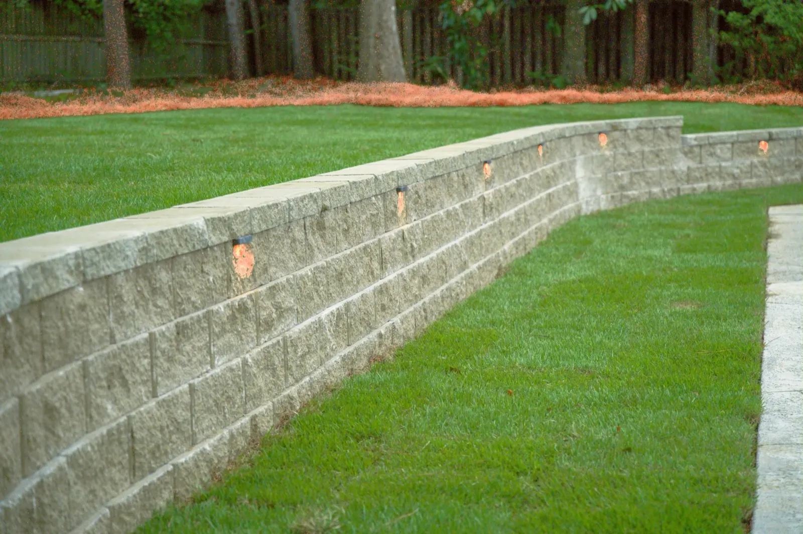 Curved block retaining wall with a capped cap course and low-voltage lighting along a backyard lawn in Baldwin County, Alabama